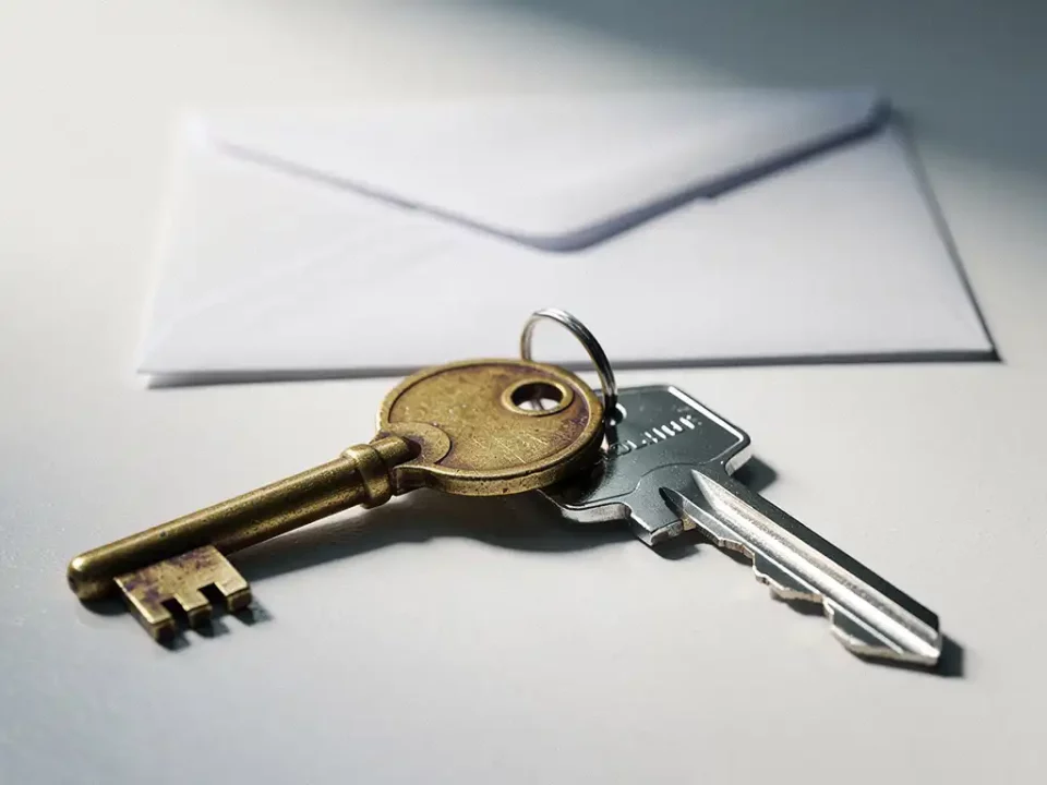 Worn brass master key beside a shiny modern key on a white desk, with a sealed envelope nearby, shot in warm gold and silver tones.