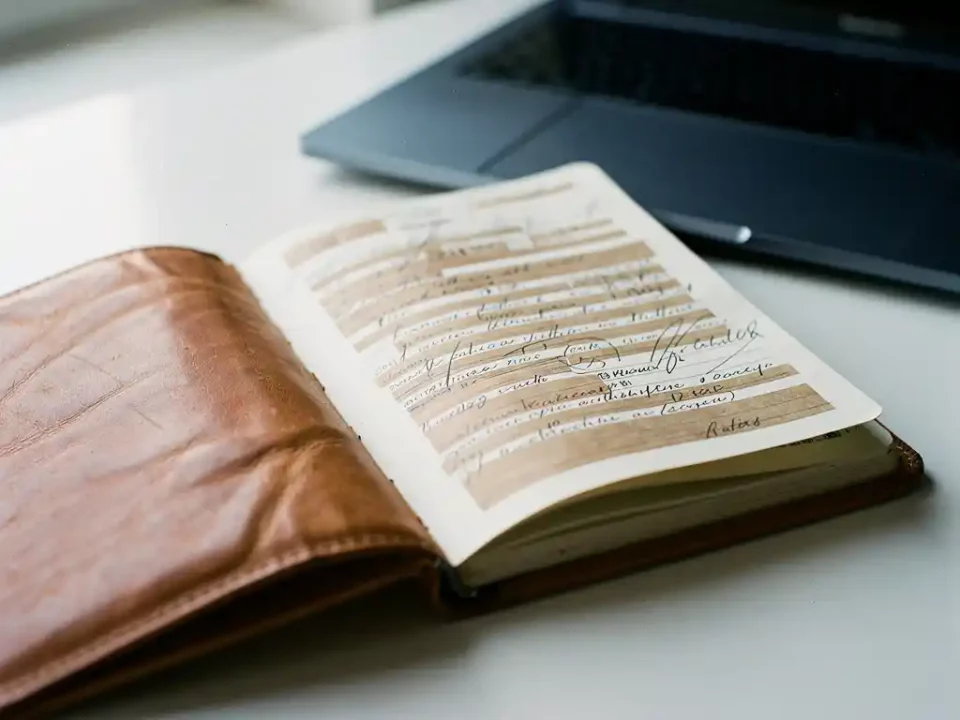 Worn leather address book open on a white desk with crossed-out and highlighted entries, laptop visible in background.