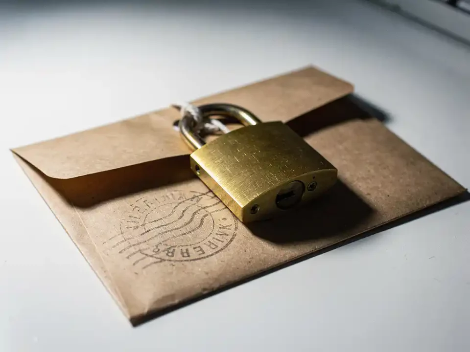 Sealed envelope secured with a brass padlock on a white desk, postal stamp impression visible, natural side lighting with warm amber tones.